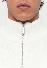 White zippered sweater with a high ribbed collar and textured knit fabric. Silver zipper hardware runs down the front.