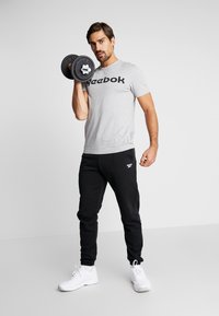 Grå T-skjorte med "Reebok"-logo, svarte treningsbukser, holder en svart manual, hvite treningssko, nøytral bakgrunn.