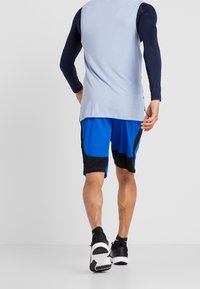 Man i blå och svarta sportshorts, en ljusblå ärmlös topp över en marinblå långärmad skjorta, och svarta träningsskor, som går bort.