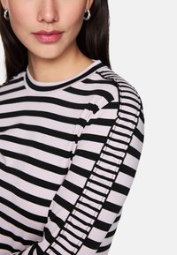 Frau in einem schwarz-weißen, horizontal gestreiften Oberteil mit Rüschendetails an der Schulter und den Ärmeln, Lippen mit subtiler rosafarbener Lippenfarbe.