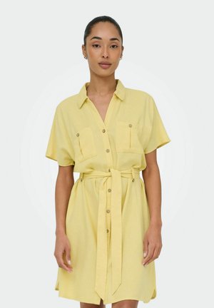 Femme portant une robe jaune à manches courtes avec des poches poitrine et une ceinture à la taille, debout devant un fond blanc.