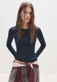 Hosszú ujjú, sötétkék, rugalmas anyagból készült felső, kerek nyakkivágással és slim fit szabással. Rózsaszín és bordó színű, megkötős derékdísz kíséretében.