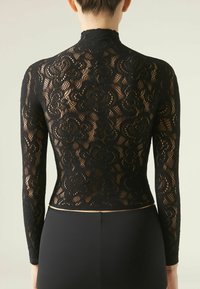 Top in pizzo nero con maniche lunghe, colletto alto e motivo floreale. Il design è trasparente con una base nera solida. Accentuato con una cintura dorata.