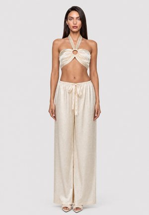 Kobieta nosząca kremowy crop top typu halter z centralnym pierścieniem oraz pasujące szerokie spodnie wiązane w talii, stojąca na neutralnym tle.