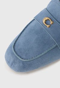 Μπλε μοκασίνι από σουέτ με στρογγυλεμένη μύτη, που διαθέτει αντίθετη πράσινη ραφή και χρυσό έμβλημα ως διακοσμητικό στο επάνω λουράκι.