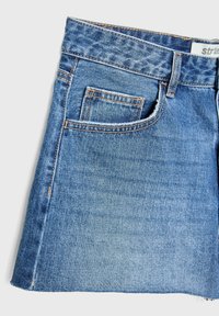 Jupe en denim bleu avec passants de ceinture à l'avant et une petite poche avant cousue à l'intérieur d'une poche plus grande sur un fond clair.