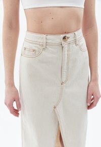 Beige denimhame, où l'avant est fendu, fermeture avec un bouton en haut et coutures visibles. Elle a des poches latérales et une coupe structurée.