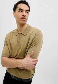 Bežinis megztas polo marškinėliai su apykakle, trumpomis rankovėmis ir trimis sagomis. Modelis su sukryžiuotomis rankomis, vilkintis juodą diržą.