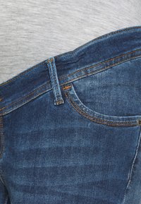 Jeans de ganga azul-escuro com costuras laranja. Apresenta um bolso frontal e uma cintura ligeiramente curva. A textura é suave e resistente.