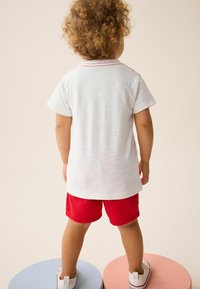 Kortærmet hvid T-shirt med rød syning, parret med røde shorts. Barnet står på runde pastelfarvede platforme, vendt væk.