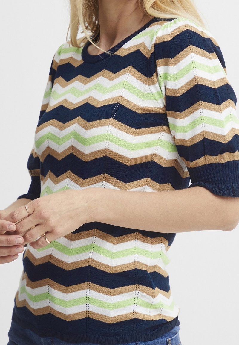 Femme portant un haut en maille à manches courtes avec des rayures en zigzag bleu marine, marron, blanc et vert, ajustant ses mains au niveau de la taille.