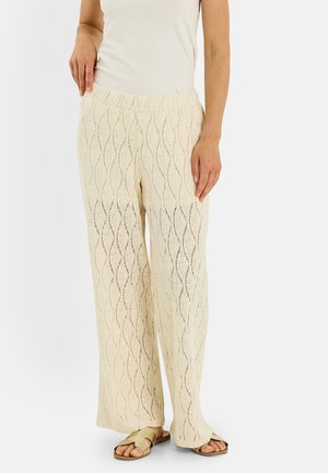 Mujer con pantalones anchos de ganchillo color crema, sandalias beige y una camiseta blanca sin mangas, de pie frente a un fondo liso.