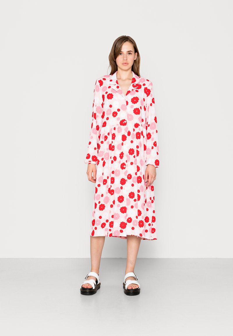 Vestido de manga longa, estampado com flores em tons de rosa e vermelho. Com comprimento até ao joelho, colarinho e corte solto, combinado com sandálias brancas e solas pretas.