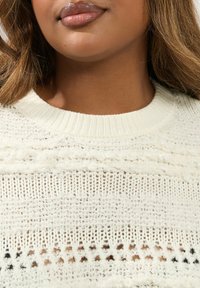 Biały sweter dzianinowy z teksturowanym wzorem, z ribbowanym dekoltem i detalami w ażurowym stylu. Miękka i lekka tkanina.