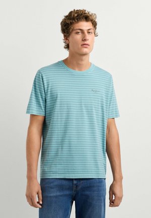 Junger Mann mit lockigem Haar, der ein hellblau gestreiftes Kurzarmhemd und blaue Jeans trägt, steht vor einem schlichten hellen Hintergrund.