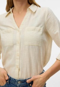 Femme portant une chemise crème à boutons avec deux poches poitrine et des manches trois-quarts, rentrée dans un jean bleu.