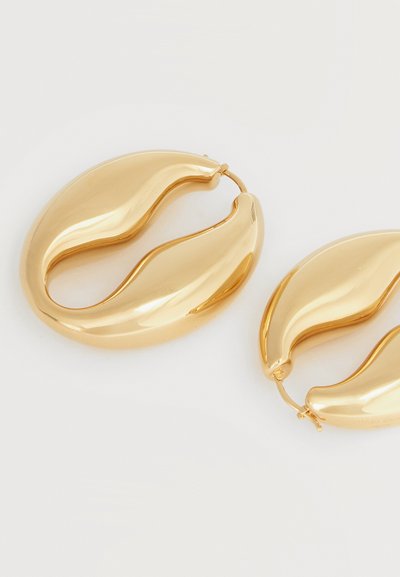 Par af skinnende guldfarvede øreringe med en bølget, ujævn oval form og låsebeslag på hvid baggrund.