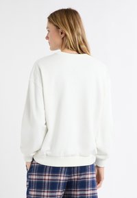 Jasnobeżowa bluza z długimi rękawami i ściągaczami. Posiada okrągły dekolt i luźny krój. Połączona z kratkowatymi spodniami do piżamy.