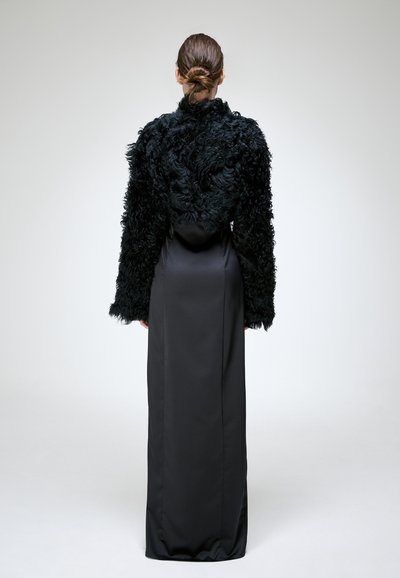 Personne debout de dos portant une longue robe noire en satin et une veste en fourrure noire texturée, les cheveux en chignon bas sur un fond uni.