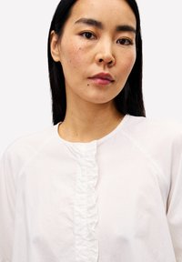 Frau mit langen, dunklen Haaren, die eine weiße Bluse mit Rüschendetails entlang der Vorderseite trägt und ruhig in die Kamera schaut.