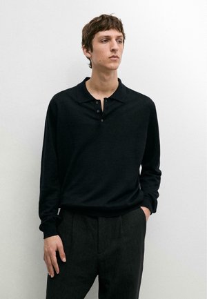 Junger Mann steht vor weißer Wand, trägt schwarzes langärmeliges Poloshirt und dunkelgraue Hose, Hand in der Tasche, schaut zur Seite.