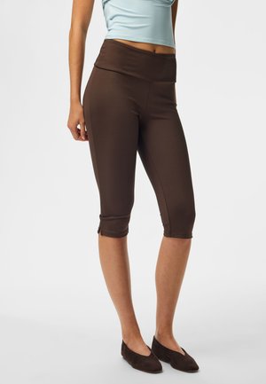 Kvinde iført brune capri-leggings og mørkebrune flade sko, står med afslappede arme, beskåret ved taljen og under knæene.
