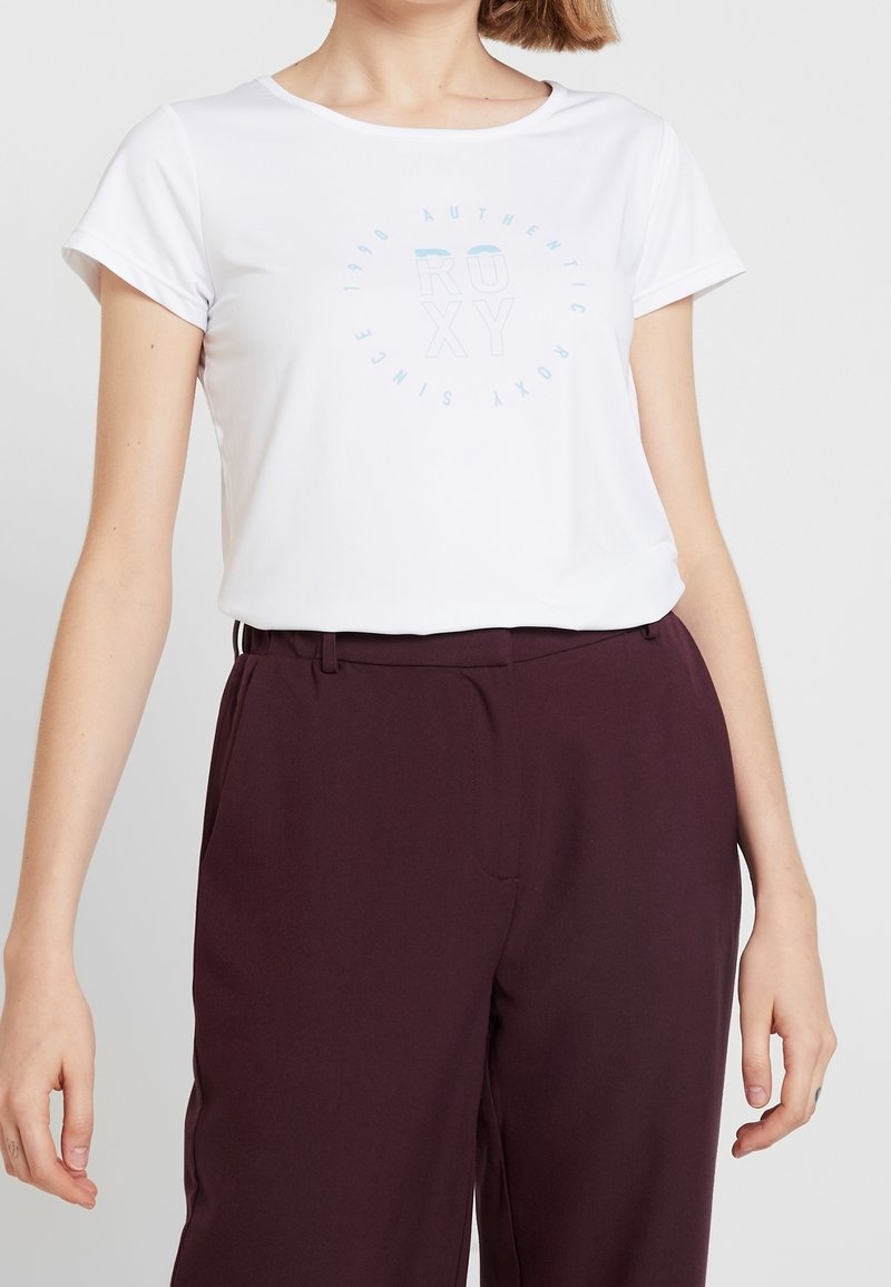 T-shirt blanc à manches courtes avec un graphique circulaire bleu clair et le texte "ROXY" ; associé à un pantalon taille haute bordeaux foncé.