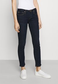 Jeans de denim azul oscuro con corte ajustado, con costuras doradas, bolsillos frontales y un logo de la marca sutil en la cinturilla. Combinados con unas zapatillas blancas.