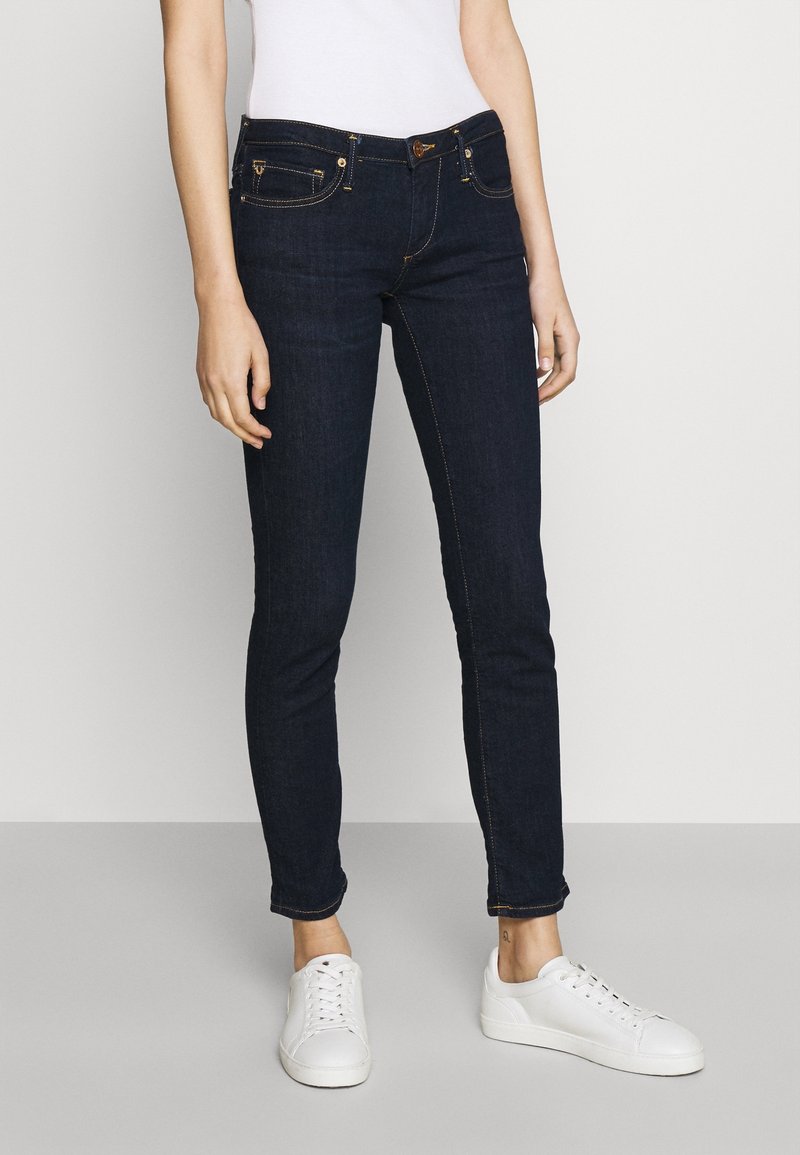 Jeans de denim azul oscuro con corte ajustado, con costuras doradas, bolsillos frontales y un logo de la marca sutil en la cinturilla. Combinados con unas zapatillas blancas.