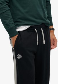 Man in zwarte sweatpants met grijze zijstrepen en trekkoorden, donkergroene sweatshirt, witte onderhemd en zwart horloge.