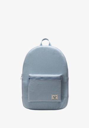 Zaino in tessuto blu chiaro con forma arrotondata, tasca frontale con zip e manico superiore. Presenta un'etichetta con logo sulla tasca frontale.