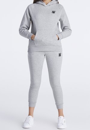 Woman wearing light grey hoodie and matching jogger pants with black logo, paired with white sneakers, standing against plain white background.