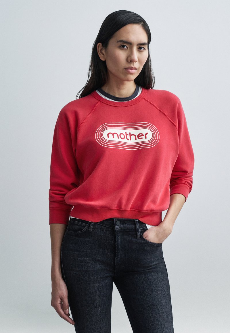 Raudonas džemperis su baltu užrašu „mother“, raglano rankovėmis ir riboto mezgimo apykakle, derinamas su tamsiais džinsais iš džinsinio audinio, klasikinio stiliaus.