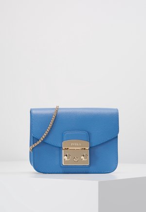 Petit sac bandoulière en cuir bleu avec fermoir en métal doré et bandoulière chaîne, placé sur une surface blanche sur un fond uni.