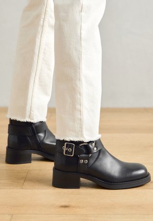 Bottes noires en cuir montant à la cheville, avec détails de boucle et de sangle argentées, portées avec un pantalon écru à ourlet brut sur un sol en bois.