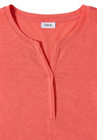Chemise rose corail à manches courtes avec un col en V fendu et une étiquette blanche portant l’inscription "Cecil" cousue à l’intérieur du col. Pas de boutons ni de motifs visibles.