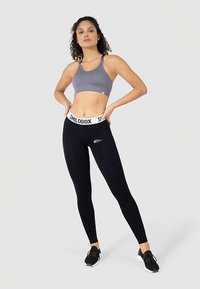 Grå sport-bh med figurnära design, kombinerad med svarta leggings som har ett vitt band och en liten logga. Modellen poserar mot en neutral bakgrund.