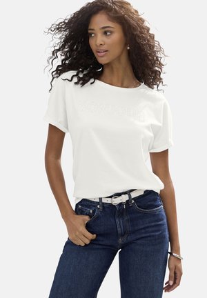 Frau mit lockigem Haar, die ein weißes "BONJOUR"-T-Shirt, dunkelblaue Jeans, weißen Gürtel und silbernes Armband trägt, steht mit einer Hand in der Tasche.