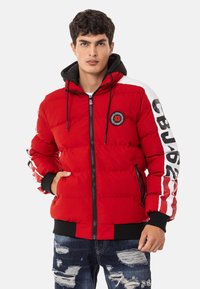 Red puffer jacket with a black hood, featuring a circular logo, zip closure, and white stripes with lettering on sleeves. Distressed denim jeans.