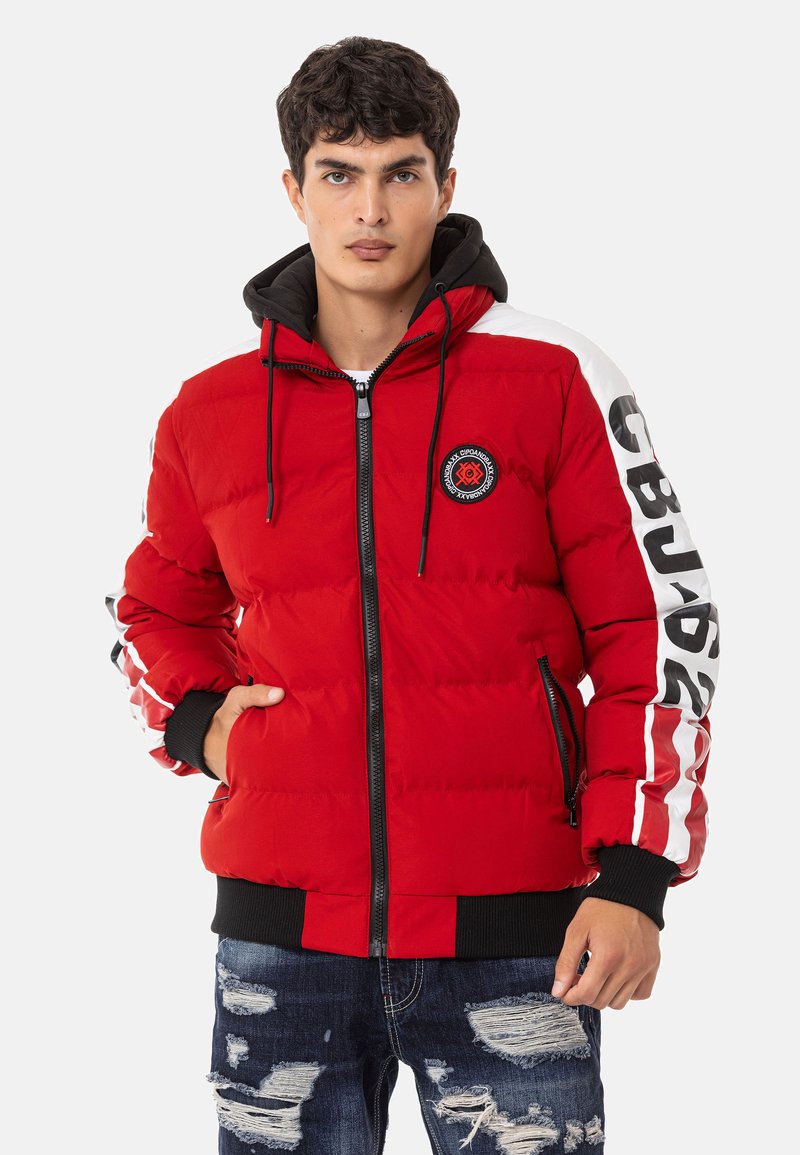 Red puffer jacket with a black hood, featuring a circular logo, zip closure, and white stripes with lettering on sleeves. Distressed denim jeans.