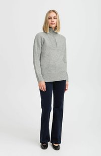 Suéter texturizado de color gris claro con cuello de media cremallera; combinado con pantalones acampanados de color azul marino oscuro; modelo de pie contra un fondo blanco.
