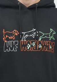 Melnā kapuce ar grafiskajiem printiem, kas attēlo krāsainus suņus oranžā, baltā un zaļā krāsā, blakus uzrakstam "HUF WORLDWIDE" burtu lielajā fontā.