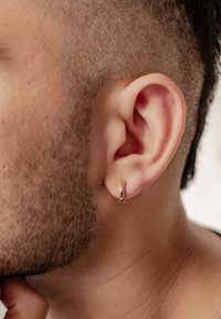 Orecchino a cerchio d'oro indossato all'orecchio sinistro, con una finitura liscia e lucida e un piccolo dettaglio di chiusura, sullo sfondo di capelli corti e ben curati.