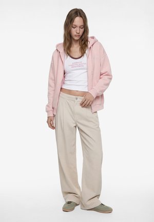 Giovane donna con felpa con cappuccio rosa, top corto bianco con testo, pantaloni beige a gamba larga e scarpe da ginnastica verdi, in piedi su sfondo bianco.