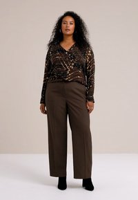 Bluză cu model maro, cu mâneci lungi, asortată cu pantaloni drepti, cu talie înaltă, de culoare maro închis și botine negre. Fundal neutru.