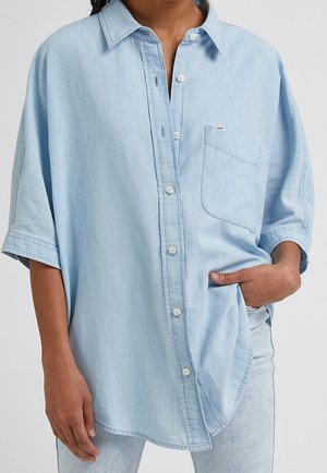 Osoba nosząca za dużą jasnoniebieską koszulę z kieszenią na piersi i jasne dżinsy, ręka w kieszeni na wysokości talii.