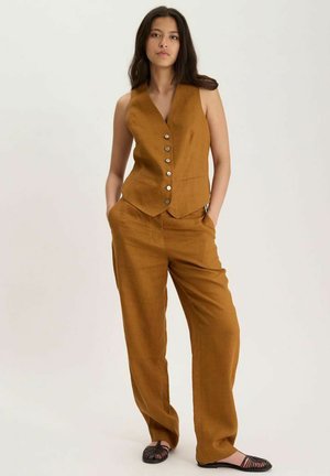 Femme debout, les mains dans les poches, portant un gilet marron boutonné, un pantalon assorti et des ballerines tissées noires sur fond uni.