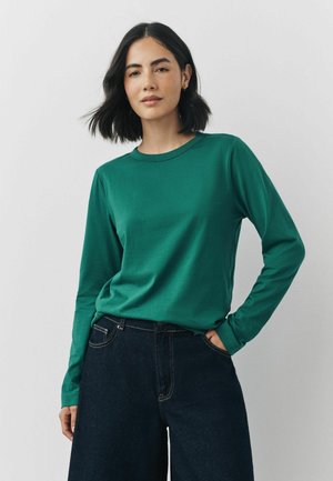 Grünes Langarm-T-Shirt aus weichem Material mit Rundhalsausschnitt, kombiniert mit dunkelblauen, weit geschnittenen Jeanshosen mit Kontrastnähten.