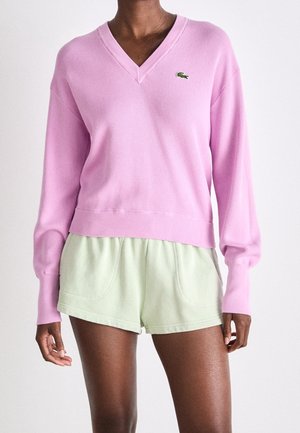 Persoon die een lichtroze V-hals trui met lange mouwen en lichtgroene casual korte broek draagt, tegen een witte achtergrond.