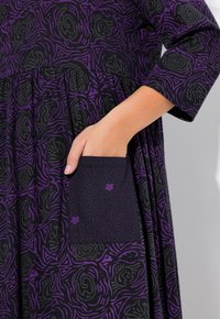 Czarna sukienka z purpurowymi abstrakcyjnymi wzorami, z marszczonym wcięciem w talii i długimi rękawami. Z boku widoczna jest solidna kieszeń w groszki.
